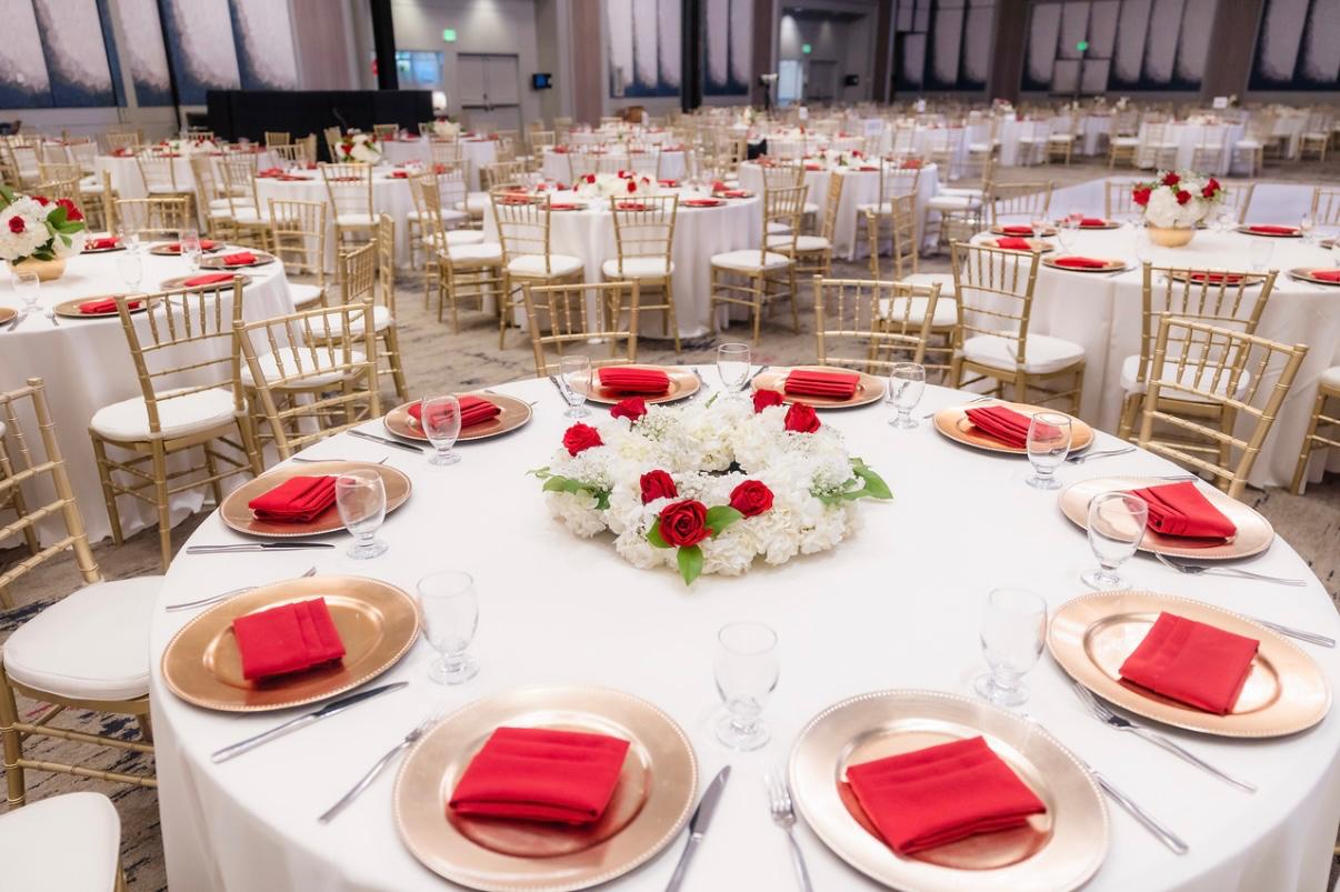 A ballroom set for an IDCA gala, with round tables in gold chargers, red napkins, and white and red floral centerpieces